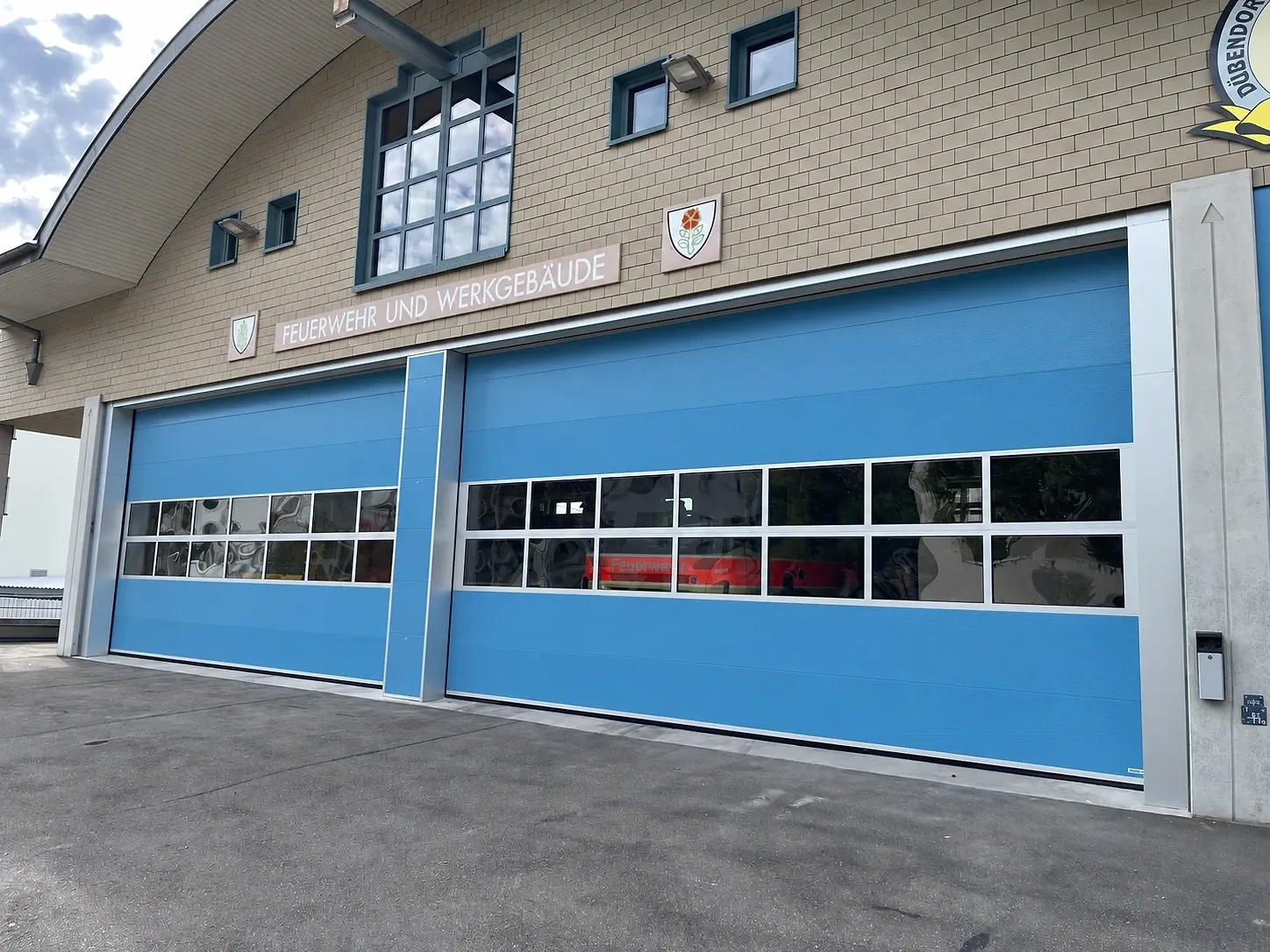 Zwei blaue Sektionaltore mit doppeltem Fensterband und blau verkleidetem Mittelpfosten am Feuerwehrgebäude Brüttisellen, hinter dem Glas ein Feuerwehrfahrzeug
